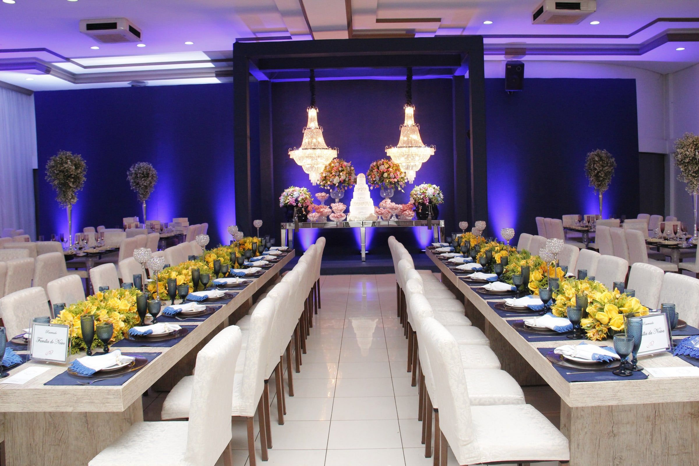 Salão de casamento com duas mesas imperiais de flores amarelas e taças azuis, corredor ao centro e lustres de cristal ao fundo