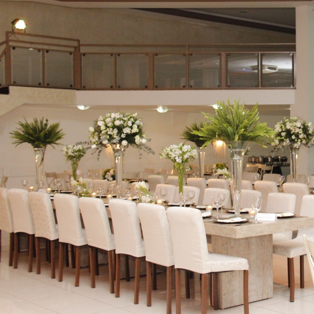 Mesa imperial de casamento com cadeiras brancas, arranjos altos de flores brancas e samambaias, e os réchauds do buffet ao fundo