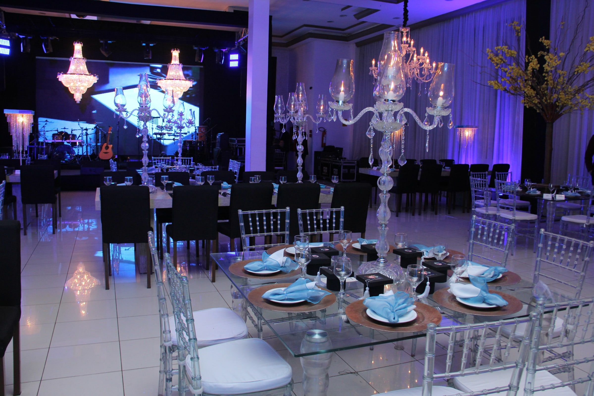 Mesa de vidro posta para baile de formatura, com candelabro de cristal, guardanapos azuis e capelos, e o palco ao fundo