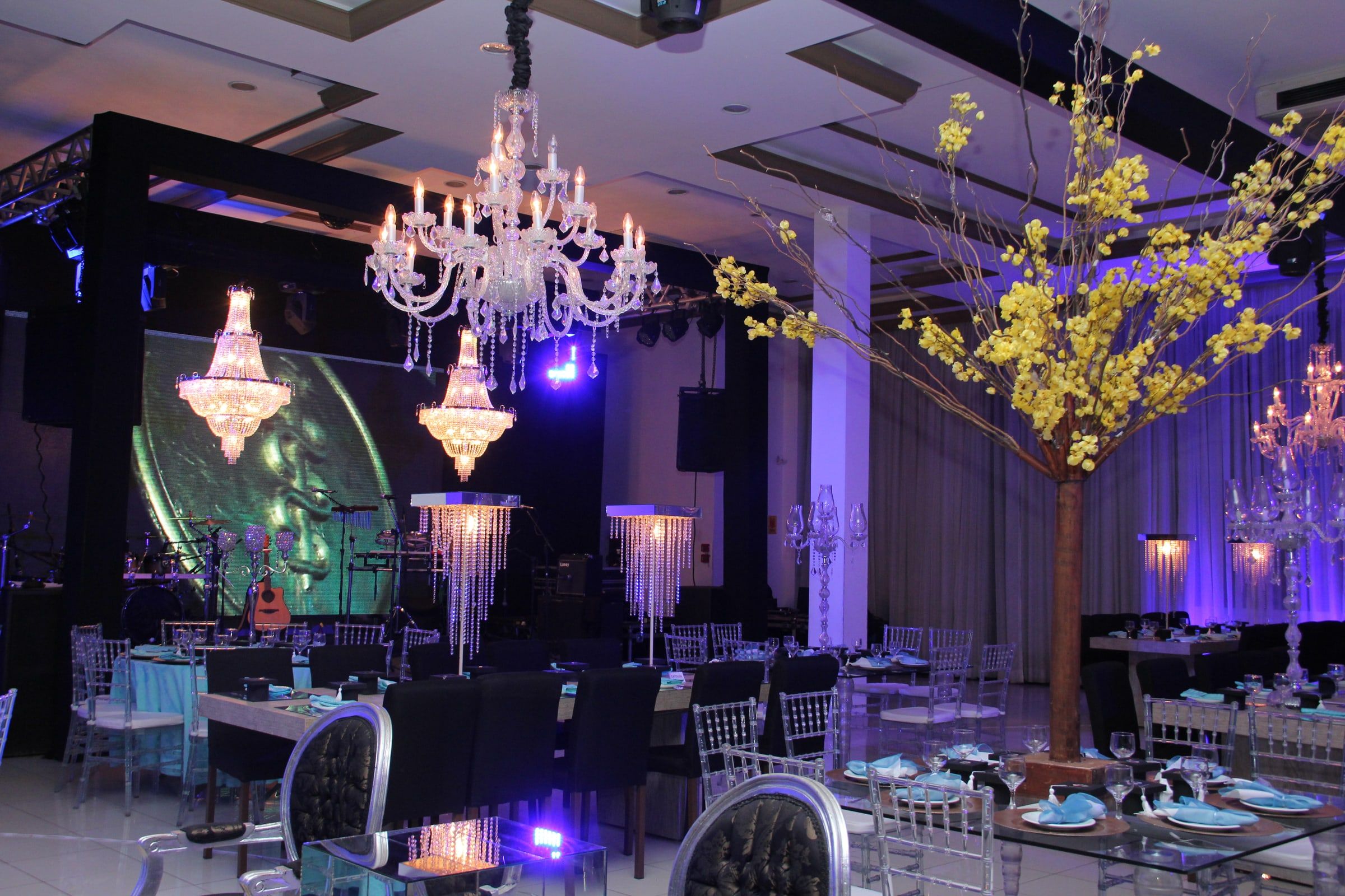 Salão de baile de formatura com lustres de cristal, árvore de flores amarelas e palco ao fundo